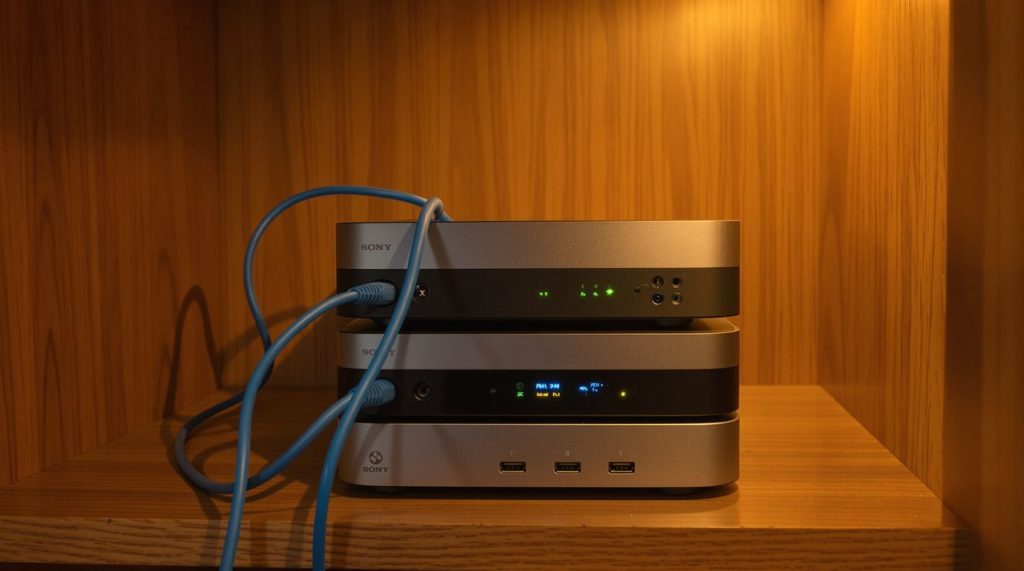 Three mini PCs stacked on a wooden shelf with blue ethernet patch cables draped between them